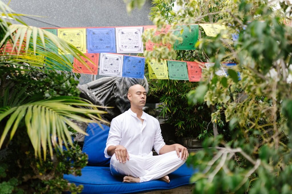 A man in white meditates outdoors in a garden, surrounded by vibrant prayer flags and greenery.