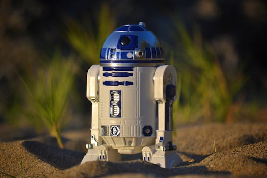 Close-up of an R2-D2 toy standing on sand, set against a green background in bright daylight.